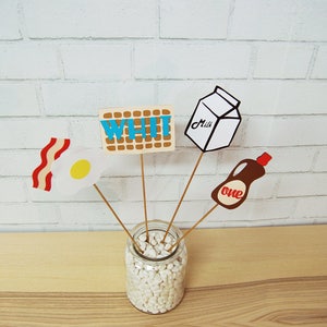 May include: A white mason jar filled with white pebbles, with four sticks holding up various breakfast-themed paper cutouts. The cutouts include a carton of milk labeled "Milk", a bottle of syrup labeled "one", a piece of bacon and an egg, and a waffle with the word "WHI" written on it.