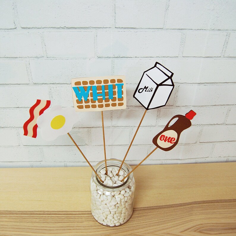 May include: A white mason jar filled with white pebbles, with four sticks holding up various breakfast-themed paper cutouts. The cutouts include a carton of milk labeled "Milk", a bottle of syrup labeled "one", a piece of bacon and an egg, and a waffle with the word "WHI" written on it.