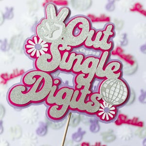Può includere: Un topper per torta di glitter rosa e argento con il testo "Out Single Digits" e un segno di pace. Il topper include anche una palla da discoteca e due fiori bianchi.