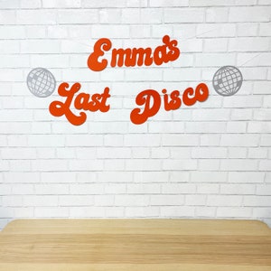 May include: Orange and white paper banner with the text "Emma's Last Disco" and silver disco ball cutouts. The banner is hanging on a white brick wall.