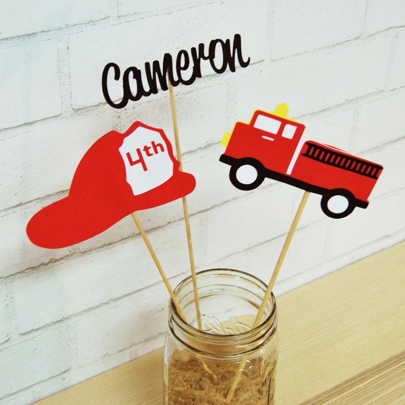 K&ouml;nnte beinhalten: Feuerwehrauto, Feuerwehrhelm und ein Schild mit dem Namen Cameron und der Zahl 4th auf Holzst&auml;bchen in einem durchsichtigen Glas.