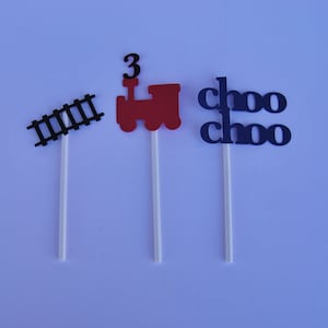 Könnte beinhalten: Drei Cupcake-Topper mit einem roten Zug mit einer schwarzen Nummer 3 oben, einem schwarzen Gleis und den Worten "choo choo" in Blau.