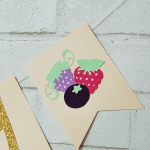 May include: A beige banner with a green, purple, and red strawberry design. The banner has a white string attached to it.