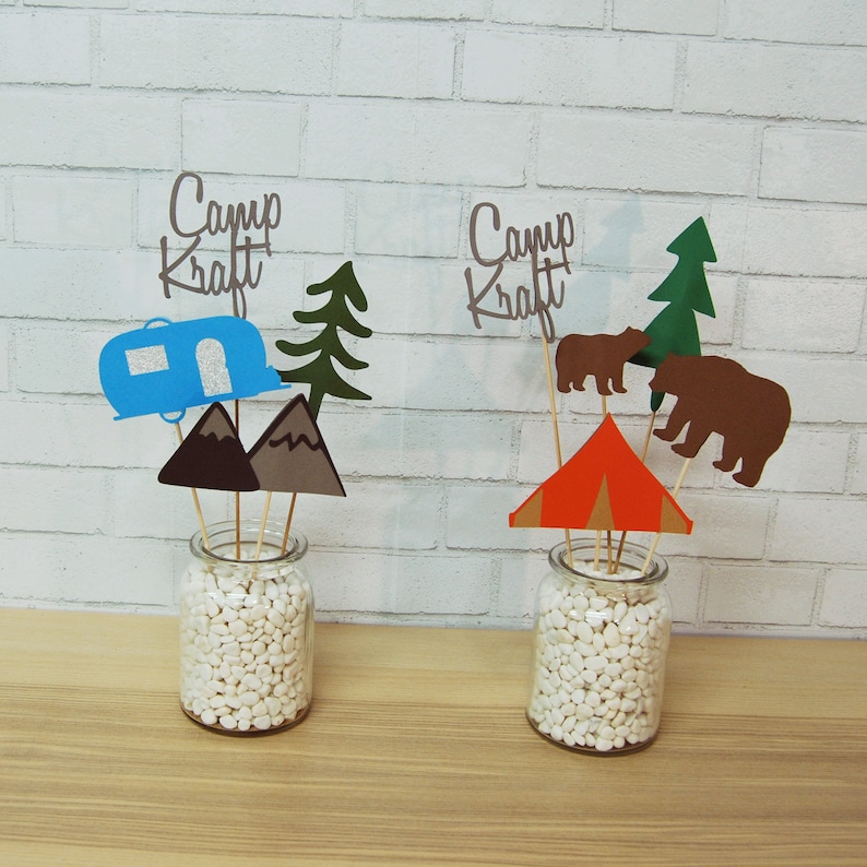 May include: Two camping themed centerpieces with white beads in glass jars. The centerpieces feature paper cutouts of a camper, mountains, a tent, trees, and bears. The centerpieces have the text "Camp Kraft" on them.