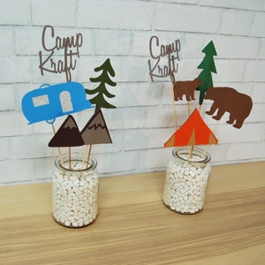 May include: Two glass jars filled with white decorative stones, each topped with a camping-themed centerpiece. The centerpieces feature paper cutouts of a camper, mountains, a pine tree, a tent, and bears. The jars are labeled "Camp Kraft".