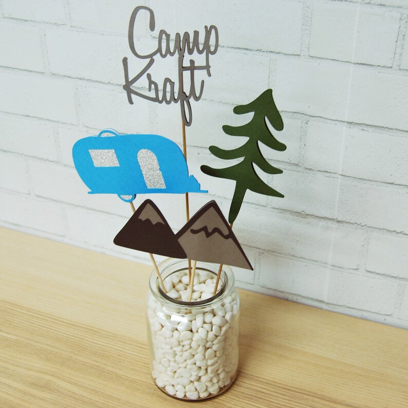 May include: A centerpiece for a camping themed party. It features a blue camper, two brown mountains, a green pine tree, and the words "Camp Kraft" in silver lettering. The decorations are arranged in a clear glass vase filled with white pebbles.