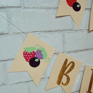 May include: A banner with a light brown background and gold glitter letters spelling "Birthday". The banner has a repeating pattern of red and white strawberries and purple blueberries with green leaves.