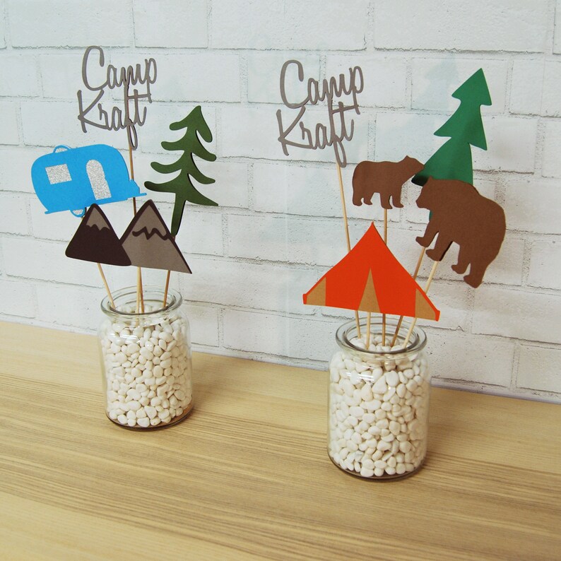 May include: Two glass jars filled with white pebbles, each with a different set of paper cutouts. The cutouts include a camper, mountains, a tree, a tent, bears, and a sign that says "Camp Kraft".