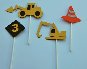 May include: Four construction themed cupcake toppers.  A yellow bulldozer, a yellow excavator, a black and yellow sign with the number 3, and an orange traffic cone.