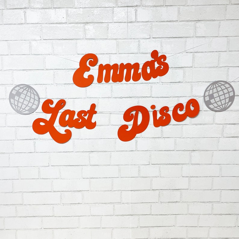 May include: Orange paper cutouts spelling "Emma's Last Disco" with silver disco ball cutouts on a white brick wall background.