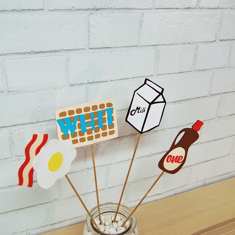 May include: A set of five paper party decorations on wooden sticks. The decorations include a carton of milk, a waffle with the word "WHI" on it, a fried egg, a strip of bacon, and a bottle of syrup with the word "one" on it.