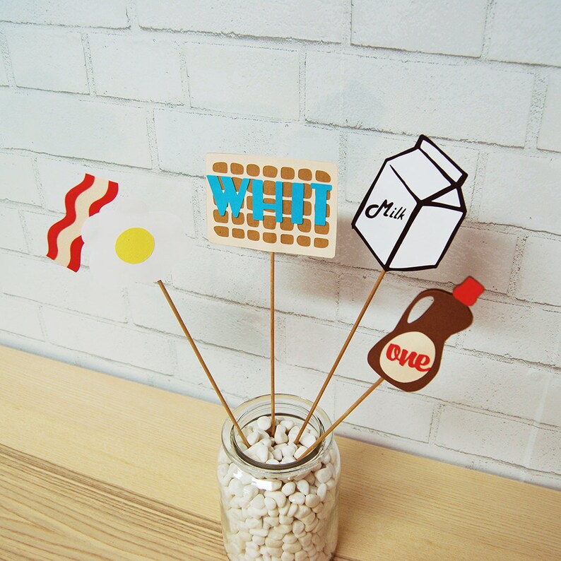 May include: A set of five paper cutouts on sticks, representing breakfast foods, in a clear glass vase filled with white pebbles. The cutouts include a waffle, bacon and egg, milk carton, and a bottle of syrup. The waffle cutout says "WHI" in blue letters.
