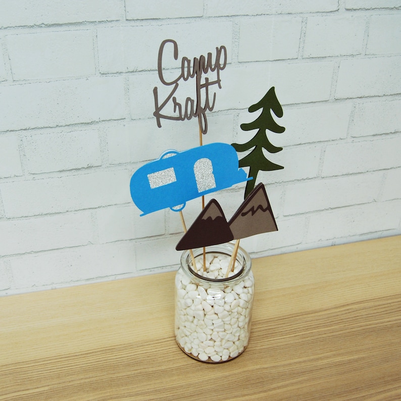 May include: A blue camper, brown mountains, and a green pine tree are arranged in a glass jar filled with white rocks. The text "Camp Kraft" is written in brown on a wooden stick.