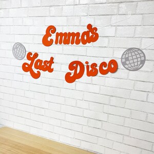 May include: Orange and white paper banner with the text "Emma's Last Disco" and two silver disco ball cutouts.