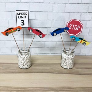 May include: A centerpiece for a race car themed party.  The centerpiece includes a red, blue, yellow and orange race car, a stop sign, and a speed limit sign that says "Speed Limit 3". The cars are attached to wooden skewers and are placed in a clear glass jar filled with white rocks.