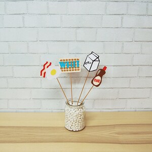 May include: A set of five paper props for a breakfast themed party. The props include a waffle, an egg, bacon, a carton of milk, and a bottle of syrup. The props are attached to wooden sticks and are arranged in a glass jar filled with white beans.