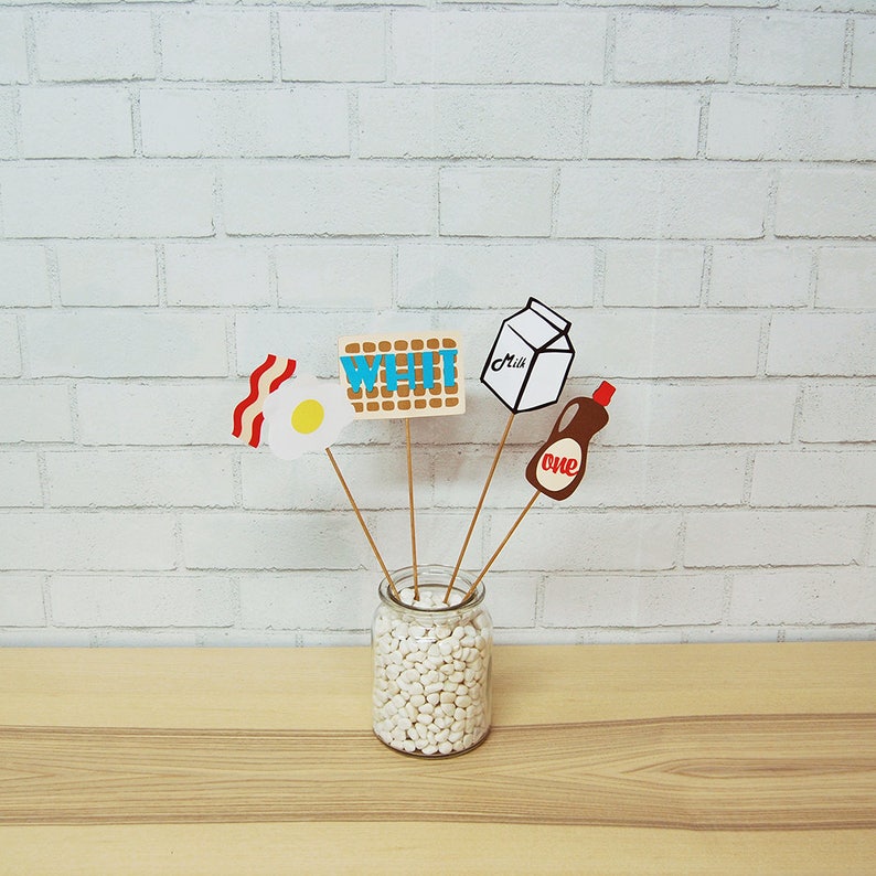 May include: A set of five paper props for a breakfast themed party. The props include a waffle, an egg, bacon, a carton of milk, and a bottle of syrup. The props are attached to wooden sticks and are arranged in a glass jar filled with white beans.