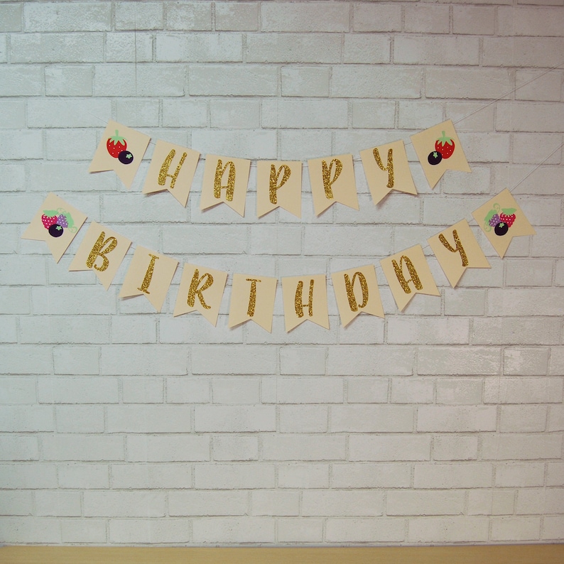 May include: A beige banner with gold glitter letters spelling "Happy Birthday" with berry and strawberry illustrations on each end.