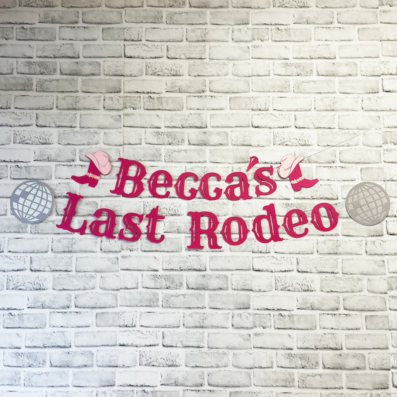 以下が含まれることがあります： 白いレンガの壁に、「Becca's Last Rodeo」と綴られたピンクと赤の紙の切り抜き。切り抜きには、カウボーイハットとブーツが含まれています。