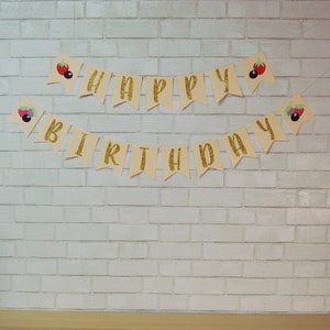 May include: A gold glitter "Happy Birthday" banner with berry and leaf illustrations. The banner is strung on a white brick wall.