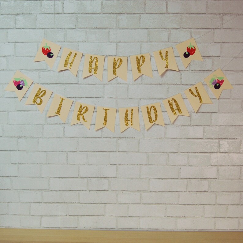 May include: A gold glitter "Happy Birthday" banner with berry and leaf illustrations. The banner is strung on a white brick wall.