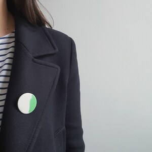 Puede incluir: Una persona lleva una chaqueta negra con una camisa a rayas blancas y verdes. Un pin circular blanco y verde está en la chaqueta.