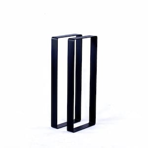 May include: Two black metal table legs with a rectangular design. The legs are made of a solid, matte black metal and are positioned against a white background. The legs are designed to support a tabletop.