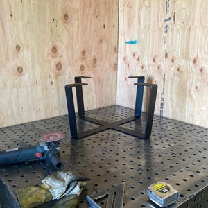 May include: A black metal table base with four legs and a square top. The base is sitting on a metal work surface with a grid pattern. There is a grinder, a pair of gloves, a metal triangle, and a tape measure on the surface.