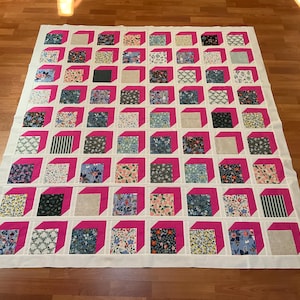May include: A handmade quilt featuring a repeating pattern of pink-framed cubes. Each cube contains a different floral or geometric fabric design. The quilt is set against a white background and is displayed on a wooden floor.