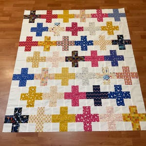 May include: A large, handmade quilt with a white background and a repeating pattern of colorful plus signs. The plus signs are in various colors and patterns, including red, blue, yellow, and patterned fabrics. The quilt is laid out on a wooden floor.