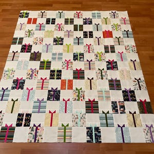 May include: A rectangular quilt featuring a grid pattern of individual gift box blocks. Each block consists of a square fabric piece in various colors and patterns, topped with a solid-colored fabric bow, all separated by white sashing strips.