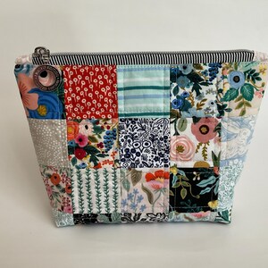 May include: A quilted patchwork pouch with a black and white striped zipper. The pouch is made of various floral and patterned fabrics in shades of red, blue, green, and pink. The pouch is a square shape.