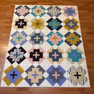 May include: A handmade quilt with a repeating pattern of colorful crosses set against a white background. The quilt features squares of various colors, including blue, pink, yellow, and black, with each square containing a cross design. The quilt is laid out on a wooden floor.