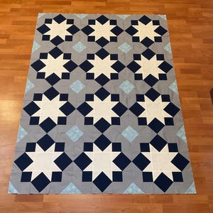 May include: A large, handmade quilt with a repeating pattern of white stars, navy blue diamonds, and light blue diamond shapes on a gray background. The quilt is laid out on a wooden floor.