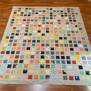 May include: A large, handmade quilt with a patchwork design. The quilt features numerous small, colorful squares in various patterns and colors, including pink, blue, yellow, and green, set against a light gray border. The quilt is laid out on a wooden floor.