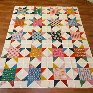 May include: A large, colorful quilt with a star pattern. The quilt features squares of white fabric interspersed with squares of patterned fabric in various colors, including pink, blue, yellow, and green. The quilt is laid out on a wooden floor.