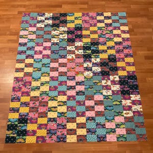 May include: A colorful patchwork quilt with a checkerboard pattern. The quilt features squares in various colors, including pink, yellow, blue, and black, each adorned with floral patterns. The quilt is laid out on a wooden floor.