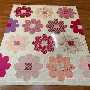 May include: A rectangular quilt featuring a repeating pattern of large, multi-petaled flower blocks in various shades of pink, magenta, and purple. Each flower has a solid square center and is set against a white background with grid lines.