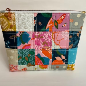 May include: A patchwork quilted zippered pouch with a variety of floral and geometric patterns in shades of pink, blue, green, orange, and white.