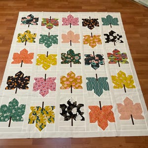May include: A handmade quilt featuring a repeating leaf pattern. Each leaf is a different color and pattern, including floral, geometric, and solid designs. The quilt is primarily white with a grid pattern.
