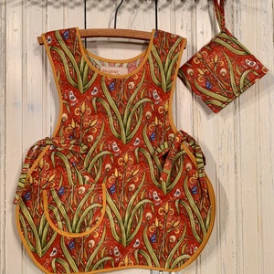 May include: A red and yellow floral print apron with a matching pocket. The apron has a yellow trim and a pocket with a blue trim. The apron is hanging on a wooden hanger.