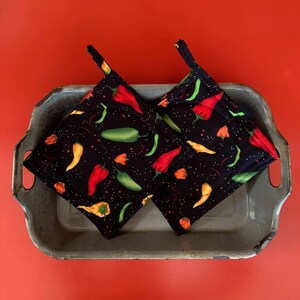 May include: Two black pot holders with a chili pepper pattern. The pot holders are red, green, and yellow on a black background. The pot holders are in a gray metal tray.