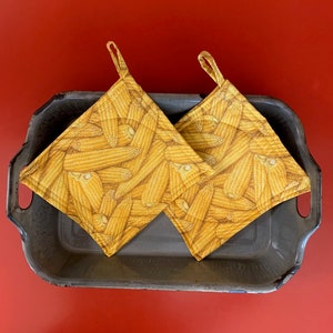 May include: Two yellow fabric pot holders with a corn on the cob pattern. The pot holders are sitting on a grey metal tray.