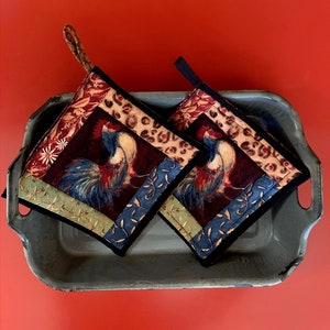 May include: Two red, blue, and green quilted pot holders with a rooster design. The pot holders are in a gray metal tray on a red background.