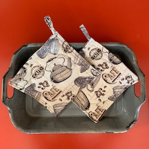 May include: Two brown and cream kitchen pot holders with a cafe theme. The pot holders have a quilted texture and feature a repeating pattern of coffee beans, coffee cups, and a teapot. The pot holders are sitting in a gray metal baking pan.