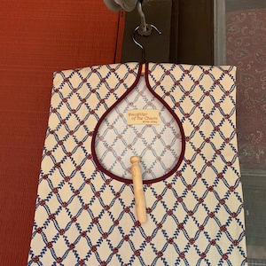 May include: A white laundry bag with a red trim and a blue and red floral pattern. The bag has a wooden dowel and a label that reads "Daughter of the Charm, Grass Valley".