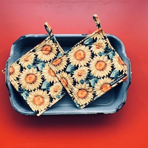 Könnte beinhalten: Zwei quadratische Topflappen mit Sonnenblumenmuster, die in einer rechteckigen Metallpfanne auf einer roten Oberfläche liegen. Die Topflappen zeigen gelbe Sonnenblumen mit orangefarbenen Zentren und grünen Blättern auf schwarzem Grund. Jeder hat eine kleine Schlaufe zum Aufhängen.