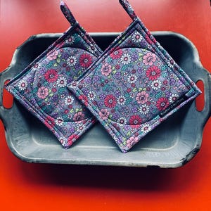 May include: Two floral patterned pot holders in pink, purple, white, and teal, set on a gray background. The pot holders have a gray border and a hanging loop. They are in a gray metal tray on a red surface.