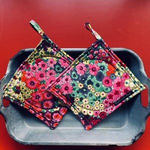 May include: Two floral print pot holders with a loop for hanging, resting on a gray metal tray against a red background. The pot holders feature a colorful pattern of pink, red, green, and yellow flowers.