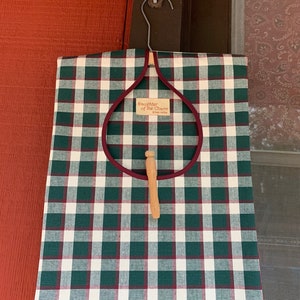 Könnte beinhalten: Eine Wäschetasche aus grünem und weißem kariertem Stoff mit burgunderfarbenem Besatz und einem Holz-Wäscheklammer. Die Tasche hat ein Etikett mit der Aufschrift "Daughter of the Charm, Green Valley".